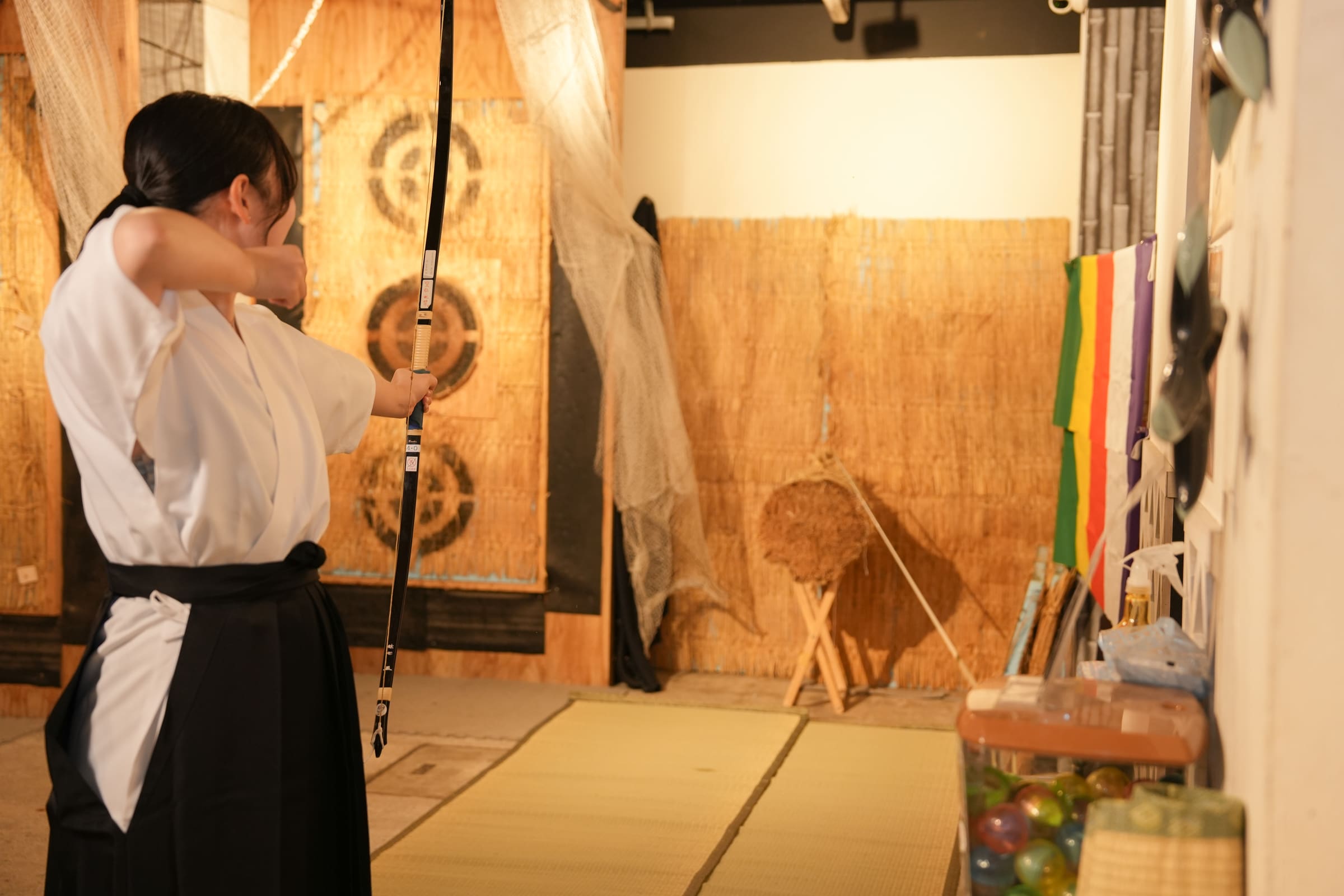 Archer in hakama aiming at a straw makiwara target across tatami mats in a kyudo practice range